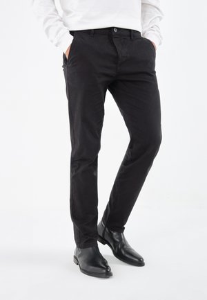 Mann trägt schwarze Slim-Fit-Hosen mit Seitentaschen, schwarze Chelsea-Stiefel aus Leder und ein weißes Langarmhemd, steht mit den Händen in den Taschen.