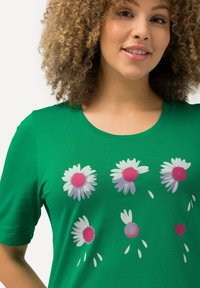 Camiseta de algodón verde con mangas cortas que presenta un diseño floral de margaritas blancas con centros rosas y pétalos dispersos, además de un detalle en forma de corazón.