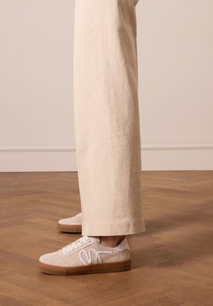 Witte suède sneakers met een tan rubberen zool, met een gestileerd logo, gedragen met losse beige broeken tegen een achtergrond van een houten vloer.