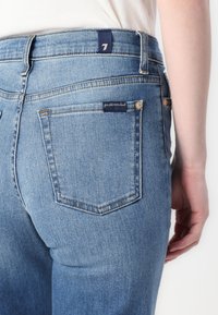Jean en denim bleu avec taille haute, design à cinq poches, surpiqûres contrastées et petite étiquette de marque sur la poche arrière.