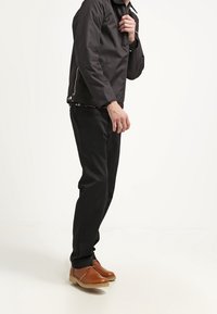 Uomo vestito con giacca nera, pantaloni neri e scarpe di pelle marrone in piedi contro uno sfondo bianco semplice.