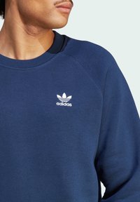 Närbild av en person som bär en blå Adidas-tröja med den vita trefoil-loggan på bröstet.