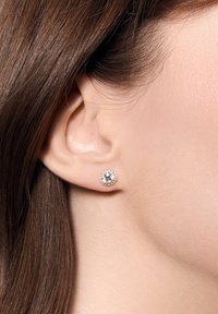 Okrągłe kolczyki-stud z centralnym przezroczystym kamieniem szlachetnym, otoczone delikatnym pierścieniem z małych przezroczystych kamieni, osadzonych w metalach w odcieniu złota.