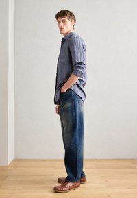 Camisa de cuadros en azul y blanco, combinada con jeans azules deslavados y mocasines marrones, vista de lado sobre un fondo neutro.