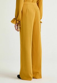 Pantaloni a gamba larga di colore giallo senape, realizzati in tessuto liscio. Presentano una vita alta, pieghe e tasche laterali. Scarpe nere visibili.