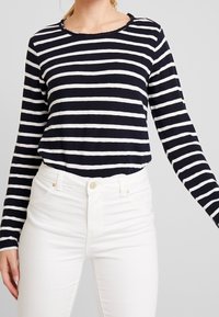 Top a maniche lunghe a righe blu e bianche, con scollatura tonda e orlo sfrangiato, abbinato a jeans bianchi a vita alta.