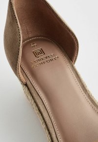 Gros plan d'une chaussure plate en daim beige avec une semelle tressée en jute et une semelle intérieure rembourrée étiquetée "Forever Comfort" en lettres dorées.