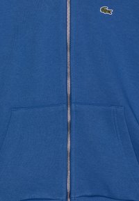 Modrá mikina na zips vyrobená z jemnej látky, s predným zipsom, dvoma prednými vreckami a malým zeleným logom na hrudi.