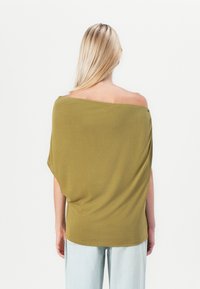 Oliven grønn av-skulder topp med en løs, drapert design. Myk, lett stoff med en avslappet passform, kombinert med lyseblå jeans.