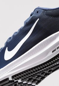 Chaussure de course en maille marine avec un swoosh Nike blanc, semelle en caoutchouc dotée d'une texture antidérapante et un rembourrage prononcé le long de la semelle intermédiaire.