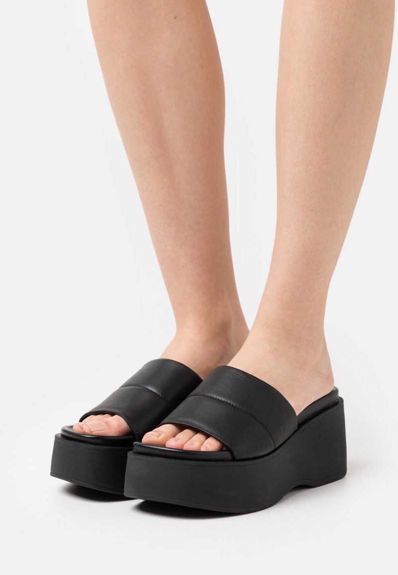 Sandalias de plataforma negras con bandas superiores anchas. Hechas de material sintético, presentan una textura suave y una forma de cuña contorneada.