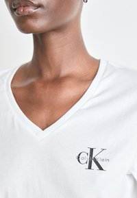 Bijela majica s V-izrezom izrađena od laganog materijala, sa malim crnim Calvin Klein logotipom na prsima, koja ističe minimalistički dizajn.