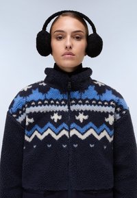 Giacca in pile con chiusura a zip, caratterizzata da un motivo geometrico blu e bianco su uno sfondo blu navy scuro. Cuffie in pelliccia nera.