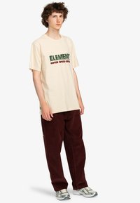 Beige t-shirt med korte ærmer med grøn og rød tekst "ELEMENT" og "OTHER" trykt på fronten. Kombineret med bordeauxfarvede fløjl bukser.