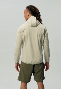 Veste à capuche beige clair avec une texture lisse et des manches longues, portée sur un short vert olive. Présente une silhouette ajustée et des poignets élastiques.