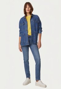 Veste en denim bleu foncé avec de grandes poches, associée à un haut en maille jaune et un jean bleu. La tenue est complétée par des baskets blanches.