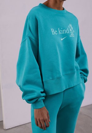 Personne portant un sweat-shirt et un pantalon bleu sarcelle, texte sur le sweat-shirt "Sois gentil" avec un petit logo Nike et un motif floral sur la poitrine.