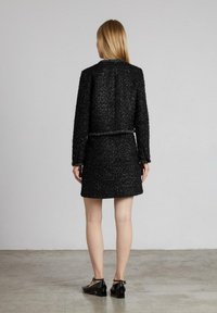 Schwarze strukturierte Tweedjacke mit passendem Rock, mit einem kragenlosen Design und einer figurbetonten Silhouette. Das Outfit wird mit schwarzen Schuhen kombiniert.