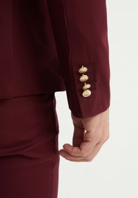 Manche de veste de costume bordeaux ornée de quatre boutons décoratifs en or et de coutures. Tissu lisse avec une coupe ajustée.