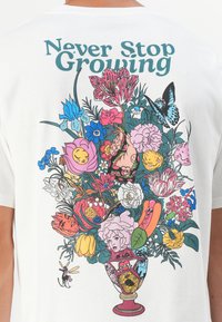 White t-shirt featuring a colorful floral graphic with various flowers, faces, and a butterfly. Text reads "Never Stop Growing."