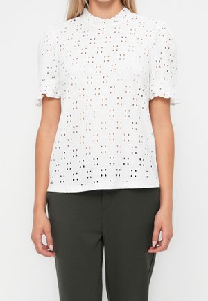 Femme portant un chemisier blanc à manches courtes avec des œillets et un col festonné ainsi qu’un pantalon vert foncé, visible des épaules jusqu’aux hanches.