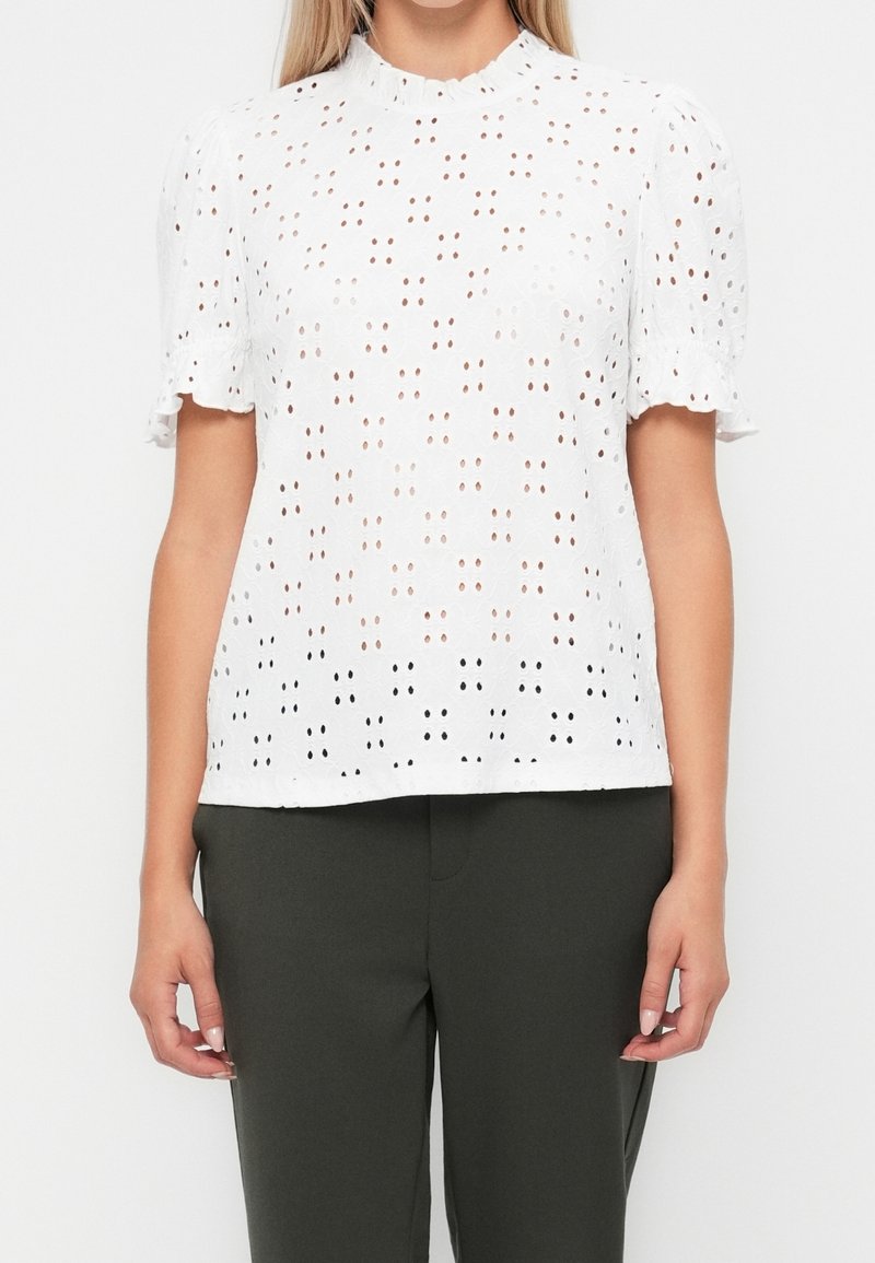 Femme portant un chemisier blanc à manches courtes avec des œillets et un col festonné ainsi qu’un pantalon vert foncé, visible des épaules jusqu’aux hanches.