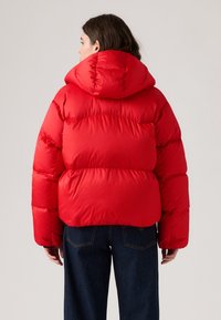 Rød pufferjakke med hætte, der har et quiltet design og en cropped silhuet. Stoffet ser glat og skinnende ud. Båret sammen med mørkeblå jeans.