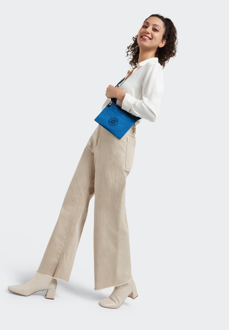 Pantaloni beige a gamba larga, blusa bianca a maniche lunghe e scarpe beige con tacco a blocco. Tiene in mano una borsa blu con zipper e un logo circolare.