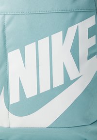 Svijetloplavi platneni ruksak s velikim bijelim Nike logotipom i glatkom teksturom, s vidljivim šavovima i bočnom džepom.