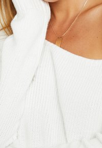 Närbild av en person som bär en off-shoulder vit stickad tröja och ett gult rektangulärt hängehalsband på en fin kedja.