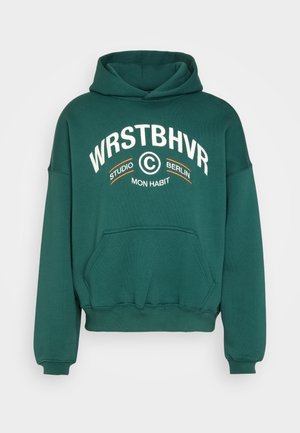 Ciemnozielona bluza z kapturem, z kieszenią z przodu i białym napisem "WRSTBHVR Studio Berlin Mon Habit" na piersi.