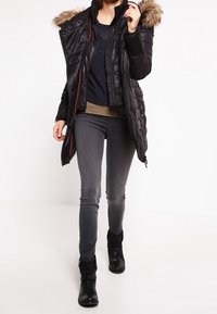 Sort pufferjakke med faux fur hætte, lynlås og knaplukning, kombineret med en marineblå trøje og grå slim-fit jeans, båret med sorte ankelstøvler.