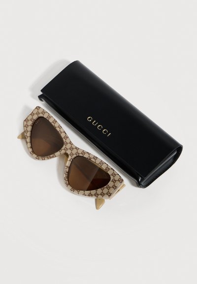 Μπεζ γυαλιά ηλίου Gucci με φακούς σε καφέ απόχρωση σε σχήμα cat-eye, δίπλα σε μαύρη θήκη Gucci με χρυσά γράμματα, σε λευκό φόντο.