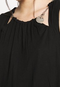 Zbliżenie na czarną bluzkę z marszczonym dekoltem, z srebrnym paskiem na ramię i okrągłymi metalicznymi zawieszkami w pobliżu ramienia.