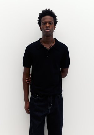 Bărbat tânăr cu păr scurt și creț, purtând un tricou polo negru cu mâneci scurte și blugi închiși la culoare, stând în picioare în fața unui perete alb simplu.