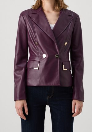 Kobieta ubrana w dopasowaną fioletową skórzaną marynarkę z złotymi guzikami i detalami przy kieszeniach, na ciemnych dżinsach, na jednolitym jasnym tle.