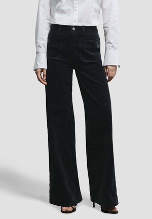 Pantalones negros de pierna ancha confeccionados en tela suave, con bolsillos delanteros y cintura alta. Combinados con una camisa blanca abotonada y tacones negros.