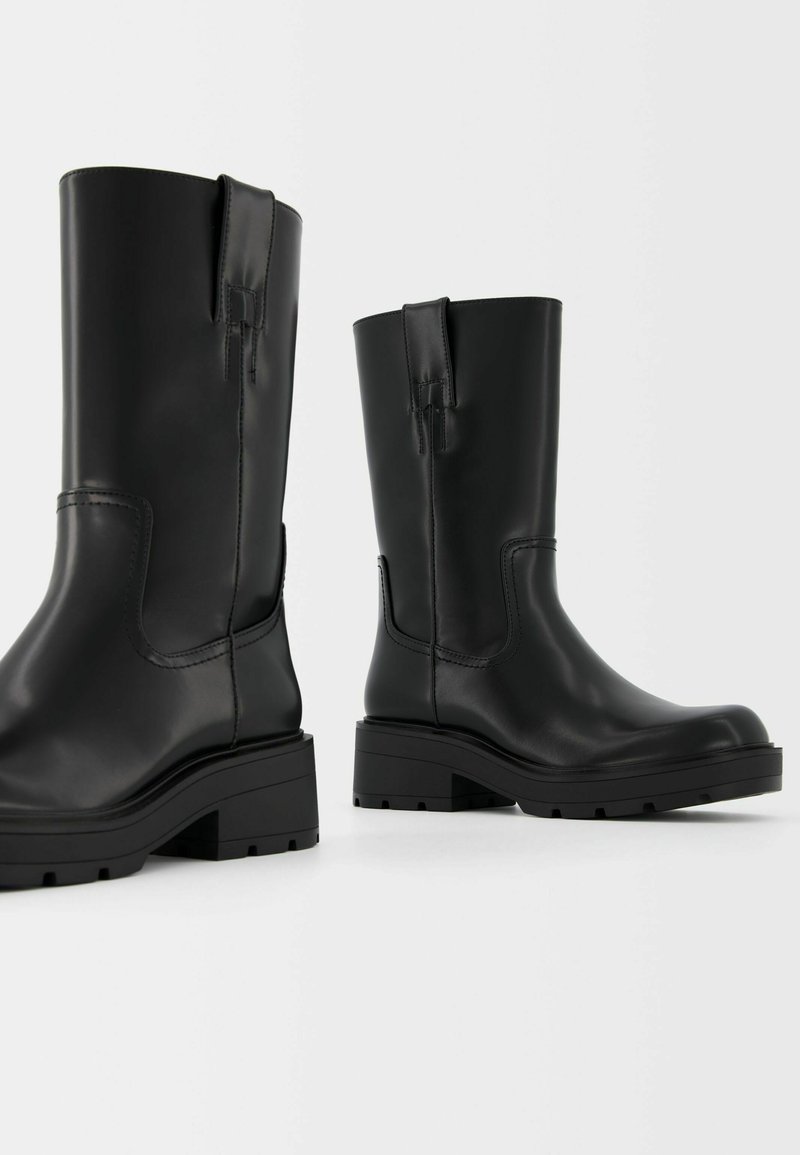 Stivali in pelle neri a metà polpaccio con linguette, suole spesse e tacco basso su uno sfondo bianco.