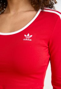 Röd långärmad topp med vit rund halsringning och randiga detaljer. Har Adidas-logotypen i vitt på bröstet. Mjuk tystkänsla.