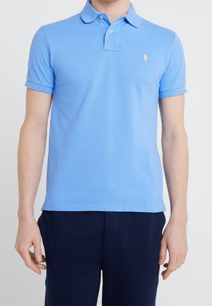 Homme portant un polo à manches courtes bleu clair avec col et boutons, associé à un pantalon marine foncé, se tenant devant un fond blanc.