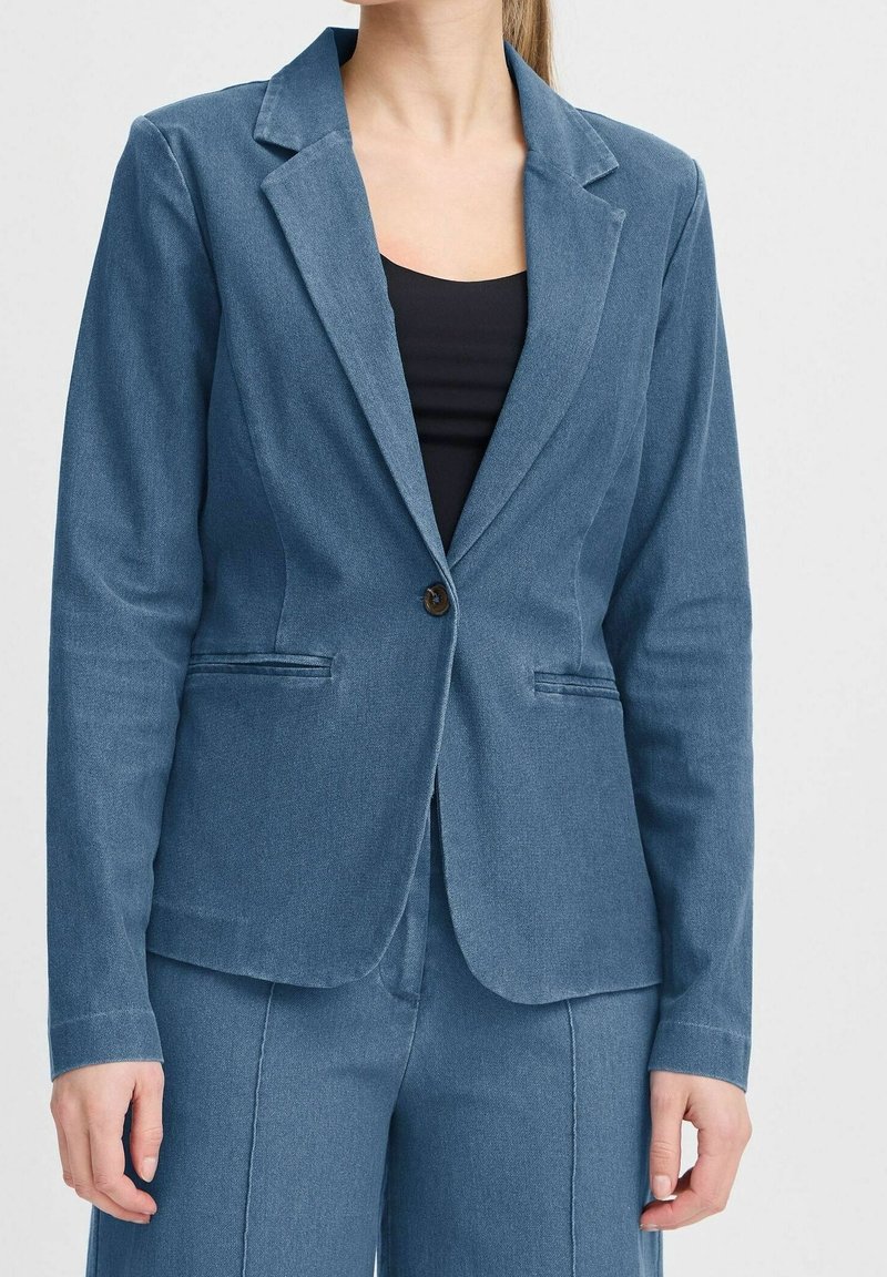 Femme portant un blazer bleu ajusté à un bouton et un pantalon bleu assorti sur un haut noir, sur un fond uni.