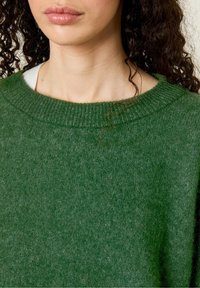 Maglione verde lavorato a maglia con un ampio scollo rotondo, texture morbida e vestibilità casual. Cuciture visibili e leggere variazioni di colore attraverso il tessuto.
