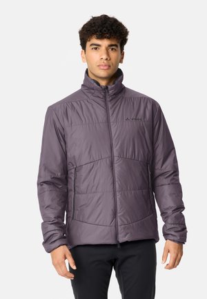 Mann mit lockigem Haar, trägt eine graue gesteppte Jacke mit Reißverschluss und schwarze Hosen, steht vor einem einfarbigen hellen Hintergrund.