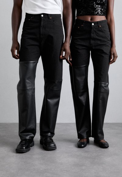 Schwarze Jeans mit Lederpaneelen an den Knien, gerader Schnitt und sichtbaren Nähdetails. Getragen mit schwarzen Schuhen.