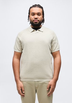 Jeune homme avec des cheveux tressés et une barbe, portant un polo à manches courtes texturé beige et un pantalon assorti, debout devant un fond blanc.