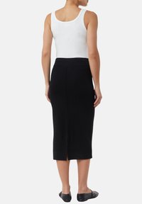 Jupe midi noire ajustée avec une fente à l'arrière, confectionnée dans un tissu lisse et extensible. Associée à un haut sans manches blanc et des ballerines noires.
