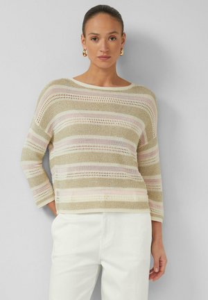 Kobieta ubrana w beżowy, jasnoróżowy i biały sweter w paski z dzianiny z rękawami do łokcia oraz białe spodnie, stojąca na jednolitym tle.