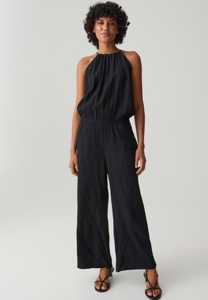 Frau mit lockigem Haar, die einen ärmellosen schwarzen Halterneck-Jumpsuit und schwarze Riemchensandalen mit Absatz trägt, stehend vor einem einfarbigen hellen Hintergrund.