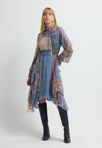 Mujer con un vestido de cuello alto, de manga larga, con un patrón de retazos y un dobladillo asimétrico, llevando botas negras hasta la rodilla, contra un fondo blanco.