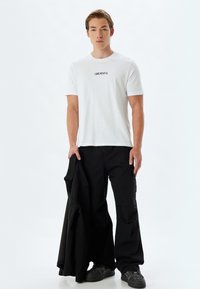 Hvid T-shirt med korte ærmer og teksten "GRATEFUL" trykt i sort. Modellen holder en sort jakke. Løse sorte cargobukser og mørke sko fuldender looket.